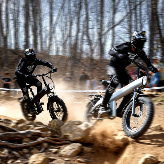 Two dirt bike riders racing on a rocky trail with spectators in the background.