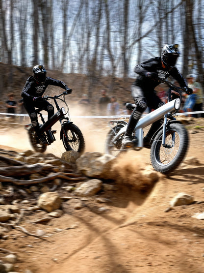 Two dirt bike riders racing on a rocky trail with trees in the background